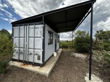 A shipping container converted into a small building with windows and a door sits on a concrete slab. It has a metal roof and is surrounded by nature.