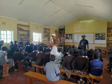 A teacher is giving a lesson to a classroom full of attentive school children sitting on benches. Bookshelves line the walls, and a projector displays images.
