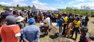 A diverse group of people gathers outdoors near a small structure, attentively listening. The event appears educational, with children and adults, some wearing uniforms, participating.