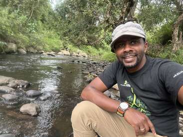 A smiling man sits by a serene, rocky river surrounded by lush greenery. He wears a hat, black t-shirt, and khaki pants, enjoying the natural outdoors.