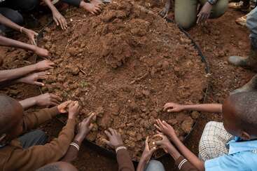 Children gather around a large circular container filled with soil, using their hands to prepare or plant, demonstrating teamwork and engagement in an outdoor gardening activity.