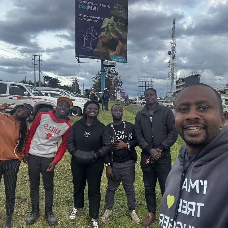 A group of six people stand together outdoors beside a busy road, smiling for a selfie. The sky is cloudy, and there are billboards and cars nearby.