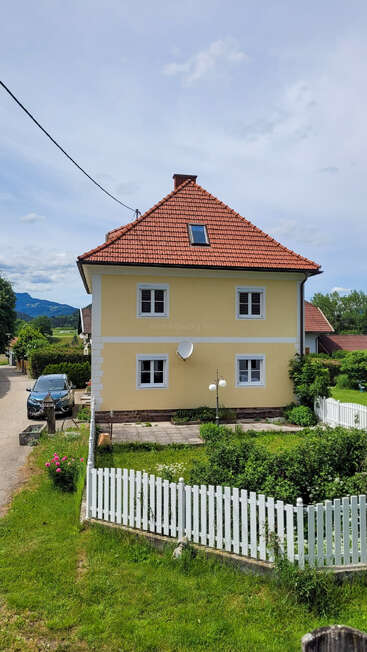 Une charmante maison jaune au toit de tuiles rouges, aux fenêtres bordées de blanc et à la clôture blanche, nichée dans un paysage rural verdoyant.