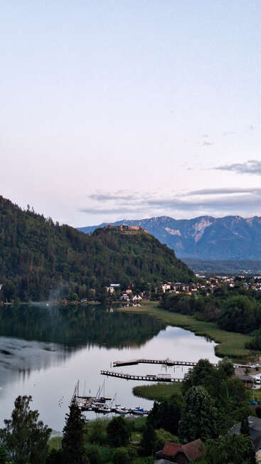 Une scène paisible au bord d'un lac au crépuscule, avec des bateaux amarrés, des collines verdoyantes, des maisons éparpillées, un château au loin et des montagnes majestueuses sous un ciel pâle. Une atmosphère paisible.