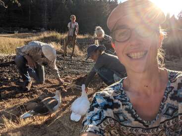 Une femme souriante prend un selfie en plein air avec des amis qui jardinent et deux canards à proximité. La lumière du soleil pénètre à flots, créant une atmosphère chaleureuse et joyeuse dans un cadre rural.