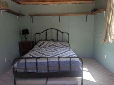 Cette chambre à coucher simple est composée d'un cadre de lit en métal, d'un édredon géométrique, d'une table de nuit en bois avec une lampe, d'étagères ouvertes, de murs vert clair et d'une fenêtre avec des rideaux rayés.