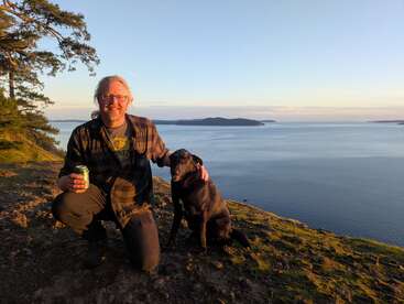 Um homem sorridente está ajoelhado ao lado de um cão preto numa encosta gramada ao pôr do sol, segurando uma lata, com vista para um mar sereno e extenso e para algumas ilhas.