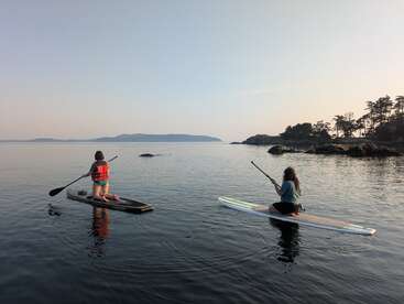 Duas pessoas estão ajoelhadas em pranchas de stand-up paddle, remando tranquilamente pelas águas calmas. O sol está se pondo, lançando uma luz suave. Árvores e ilhas são visíveis ao longe.