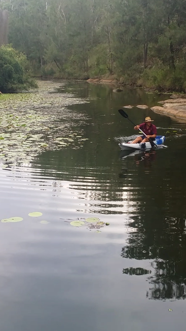 Um homem rema um caiaque em um rio com um balde na parte de trás, cercado por árvores e nenúfares, usando chapéu e camisa marrom.