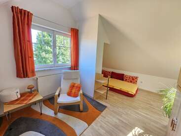 Cette chambre confortable comprend un lit simple avec des oreillers colorés, une chaise confortable, une table d'appoint, des rideaux orange, un tapis géométrique et beaucoup de lumière naturelle.