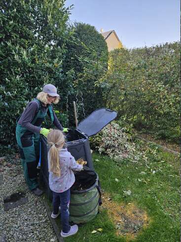Une femme et une jeune fille compostent des feuilles et des déchets de jardin dans une arrière-cour. Elles travaillent ensemble à côté de buissons verts sous un ciel bleu clair.