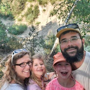 A happy family of four poses for a selfie in a beautiful outdoor setting, surrounded by greenery and smiling brightly, enjoying their adventure in nature together.