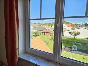 A bright, sunny day seen through a window. Outside, there are houses, a red-roofed shed, green lawns, a playground, scattered trees, and a clear blue sky.