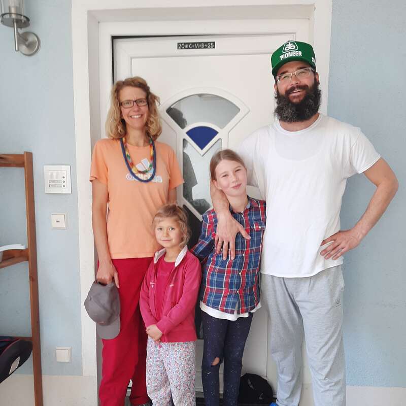 Une joyeuse famille de quatre personnes se tient devant une porte blanche. Les parents sourient fièrement à côté de leurs deux filles, habillées de façon décontractée, qui profitent d'un moment de détente ensemble.