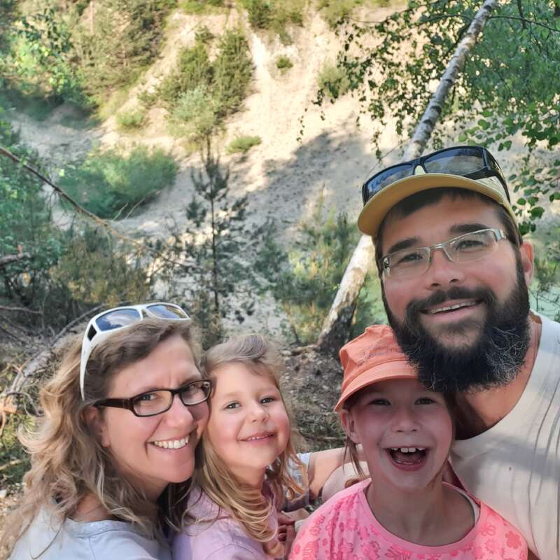 A happy family of four poses for a selfie in a beautiful outdoor setting, surrounded by greenery and smiling brightly, enjoying their adventure in nature together.