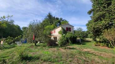 Une charmante maison à deux étages avec des volets rouges se dresse au milieu d'une végétation luxuriante et d'arbres. La cour spacieuse comporte des zones ensoleillées, quelques plantes sauvages et des meubles d'extérieur éparpillés.