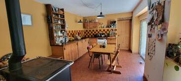 Cette cuisine confortable présente des murs jaunes, des armoires en bois chaud, un sol carrelé, des étagères éclectiques, une table de salle à manger centrale avec des chaises, et une lumière naturelle brillante provenant des portes coulissantes.