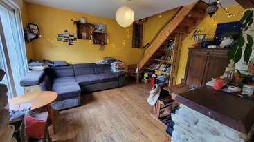 Un salon confortable avec des murs jaunes, un grand canapé sectionnel gris, des planchers en bois, des bibliothèques, des escaliers, des guirlandes lumineuses, des plantes et des objets personnels éparpillés, créant une atmosphère chaleureuse.