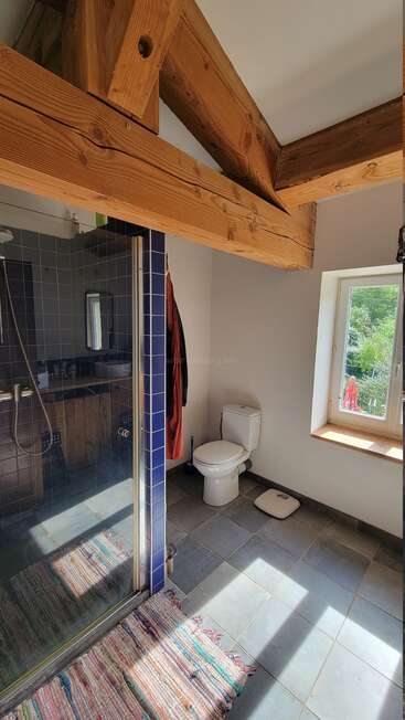 Cette salle de bains lumineuse comprend des poutres en bois apparentes, une douche en verre avec des carreaux bleus, des toilettes, une fenêtre donnant sur la verdure, une balance et un tapis à rayures sur le sol carrelé.