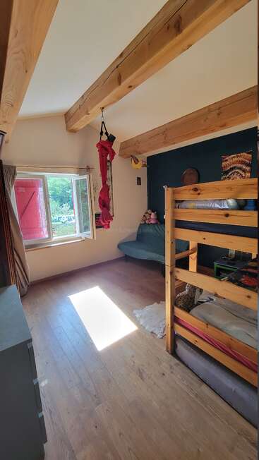 La chambre est dotée de planchers en bois, d'un lit superposé, de poutres apparentes, d'une fenêtre ouverte, d'un hamac rouge suspendu, d'un mur d'accent bleu, d'un canapé et de jouets en peluche - une atmosphère lumineuse et chaleureuse.