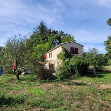 Ein charmantes Haus auf dem Land mit roten Fensterläden ist umgeben von üppigem Grün, Bäumen und bewachsenem Gras unter einem hellblauen, leicht bewölkten Himmel. Friedliche ländliche Umgebung.