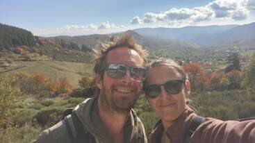 Un homme et une femme souriants portant des lunettes de soleil prennent un selfie en plein air. Derrière eux, de belles collines, des arbres et des montagnes s'étendent sous un ciel partiellement nuageux.