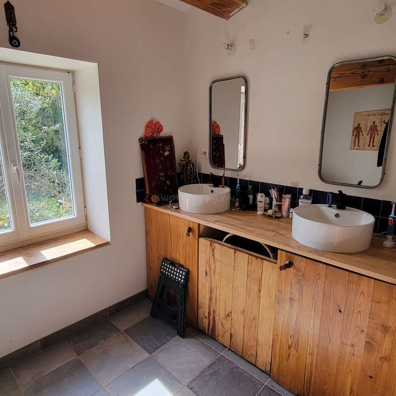 Une salle de bain lumineuse avec une grande fenêtre, deux lavabos sur un meuble en bois, deux miroirs, des articles de toilette, du carrelage et un petit tabouret pliant sur le sol.