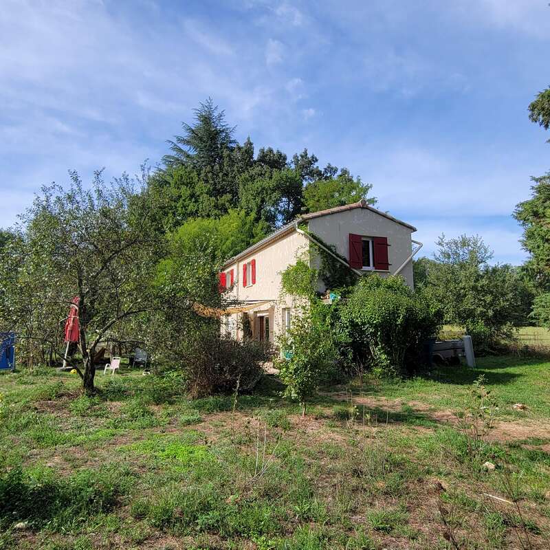 Une charmante maison de campagne aux volets rouges est entourée d'une végétation luxuriante, d'arbres et d'herbes folles sous un ciel bleu clair et légèrement nuageux. Cadre rural paisible.