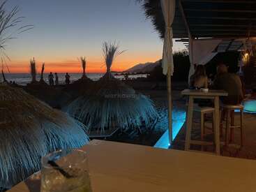 Eine wunderschöne Strandszene bei Sonnenuntergang, mit Strohsonnenschirmen, Leuten, die auf einer Terrasse chillen, einem leuchtend blauen Licht, Bergen und einem Drink auf dem Tisch.