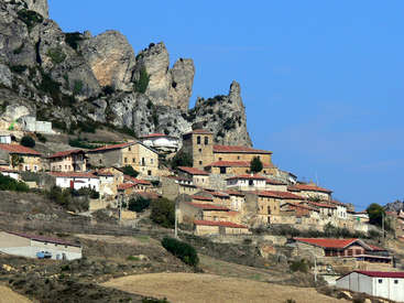 L'image représente un petit village situé au pied d'une montagne, avec un ensemble de bâtiments aux toits rouges et un ciel bleu clair.