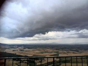 L'image représente un vaste paysage de champs et de collines sous un ciel orageux, avec une rambarde métallique au premier plan.