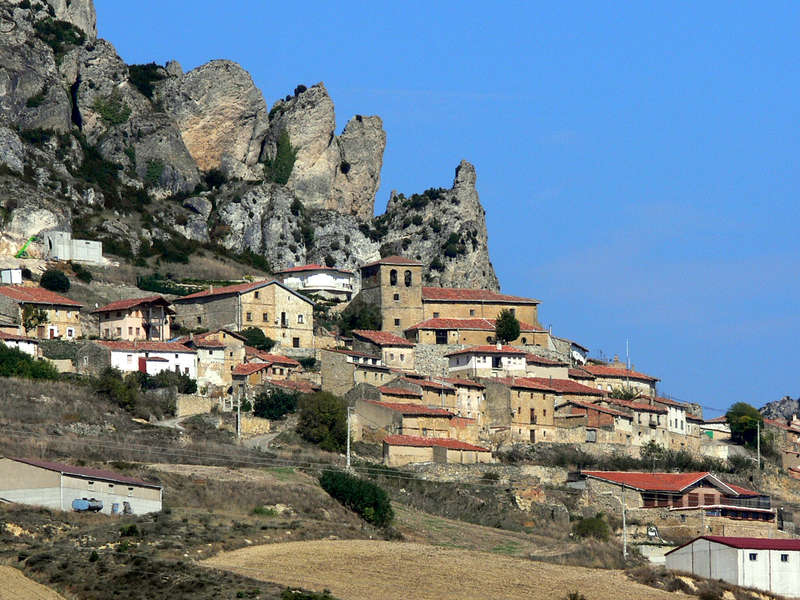 L'image représente un petit village situé au pied d'une montagne, avec un ensemble de bâtiments aux toits rouges et un ciel bleu clair.