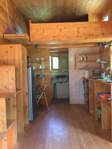 Dieses Bild zeigt das gemütliche Interieur eines kleinen Holzhauses mit offenen Regalen, einer kompakten Küche, einem Schlafbereich im Dachgeschoss, einem Kühlschrank und viel Tageslicht, das durch kleine Fenster hereinströmt.