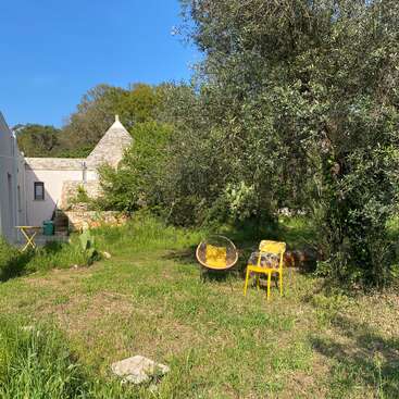 Une scène paisible dans un jardin, avec deux chaises jaunes sous un arbre, une petite table ronde près d'une maison blanche et une pelouse verdoyante sous un ciel bleu.