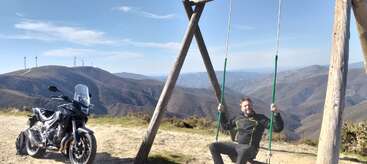 A man in motorcycle gear sits on a large wooden swing atop a mountain, with a motorcycle parked nearby and wind turbines visible in the background.