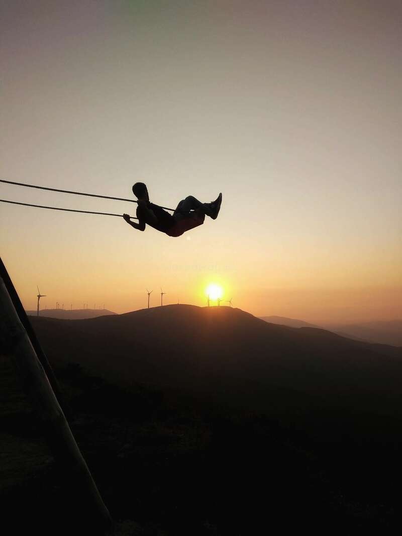A person is suspended in mid-air on a zip line, with the sun setting behind them and wind turbines visible in the distance.