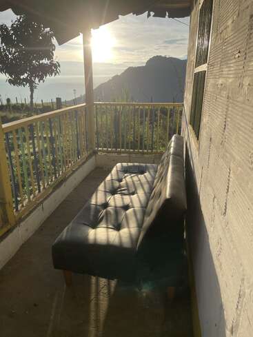 Sunlight bathes a porch with a black leather sofa, casting shadows. Scenic mountains, greenery, and a glowing horizon create a serene, peaceful outdoor retreat.