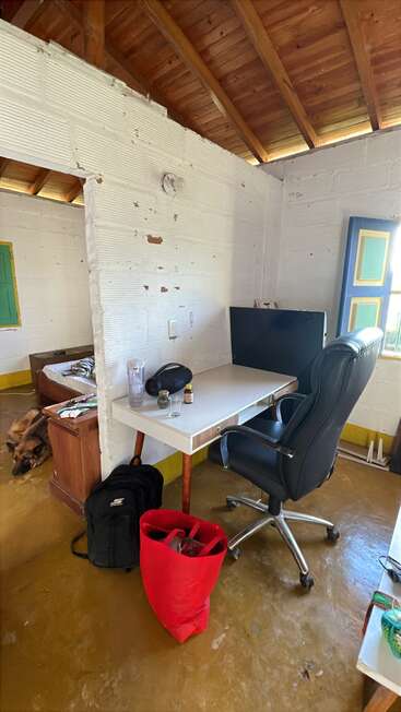 A rustic room with white brick walls and wooden ceiling features a desk, black office chair, monitor, red bag, backpack, and a dog resting by the bed.