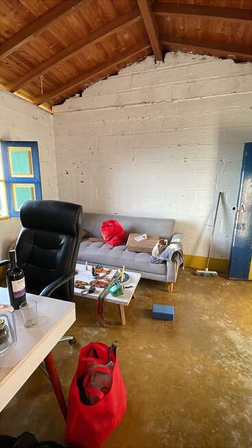 A rustic room with a wooden ceiling, messy sofa, coffee table clutter, office chair, desk with wine bottle, red bag, mop, and window with blue trim.