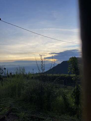 A serene landscape at sunset, featuring lush green vegetation, distant mountains, a cloudy sky with soft sunlight, and a simple wire stretching across the view. Peaceful nature.