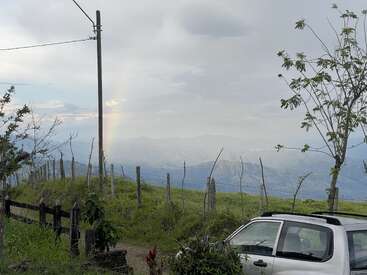 A countryside scene featuring a parked car, rustic fence, utility pole, scattered trees and plants, cloudy sky, mountains in the background, and a faint rainbow.