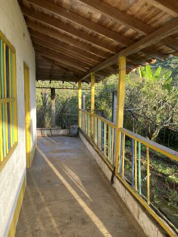 A rustic porch with a wooden roof, yellow-trimmed railings, and sunlight streaming in. Surrounded by greenery, it overlooks a peaceful garden and hills.