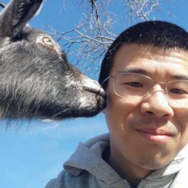 Un homme portant des lunettes et un sweat à capuche gris sourit pour un selfie en plein air, tandis qu'une chèvre lui donne des petits coups de museau ou lui renifle la joue sous un ciel bleu limpide.