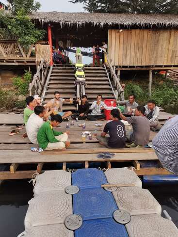 Un grupo de personas se sienta en un muelle de madera, compartiendo comida y bebida. Una rústica cabaña de madera y unas escaleras se ven al fondo, creando un ambiente relajado.
