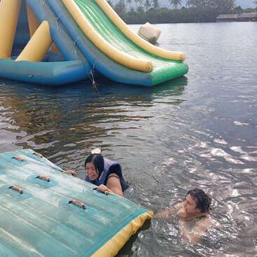 Dos personas están en el agua cerca de un gran tobogán hinchable. Una lleva chaleco salvavidas y sujeta un flotador, mientras la otra sonríe cerca. Palmeras al fondo.