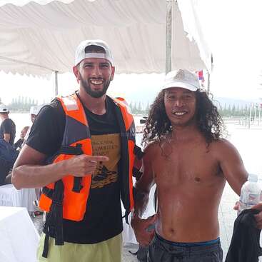 Dos hombres sonrientes posan juntos al aire libre bajo una tienda de campaña. Uno lleva chaleco salvavidas y sombrero; el otro, sin camiseta, sostiene una botella de agua; ambos parecen alegres.