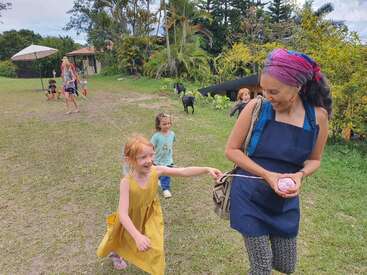 A woman with a colorful headscarf leads laughing children on a yarn trail outdoors, with goats and trees in the background, evoking joy and playfulness.