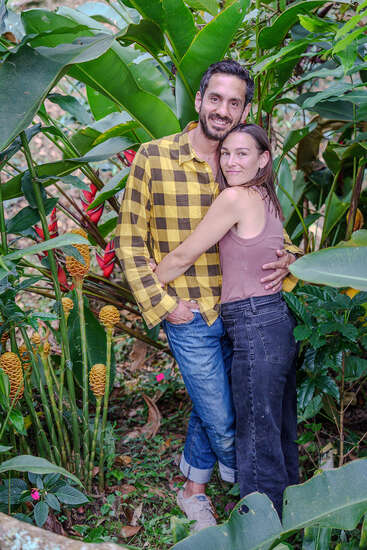 A smiling couple lovingly embraces in a lush, green tropical garden surrounded by large leaves and colorful exotic plants, radiating happiness and warmth together.