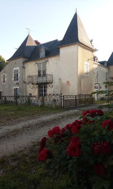 An old, charming house with pointed slate rooftops and a wrought-iron balcony stands behind a fence. Vibrant red roses bloom in the foreground. Peaceful atmosphere.