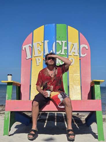 A man in a tropical shirt sits on a giant colorful chair labeled "Telchac Puerto," holding coffee, enjoying a sunny beach day with clear blue skies.